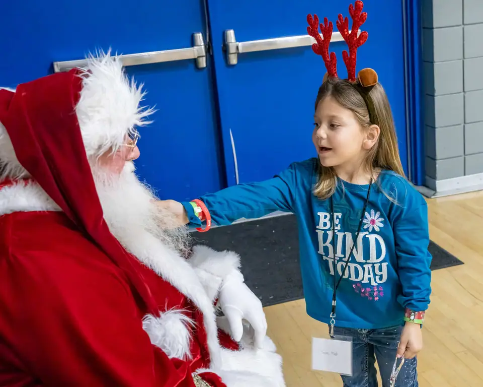A girl in reindeer antlers and a Be Kind shirt reaching out to Santa